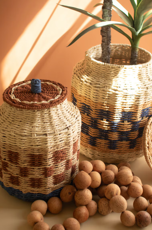 Two woven basket-style containers with lids, one small and one large, styled with a potted plant and scattered cork balls on a warm beige surface against a peach background.
