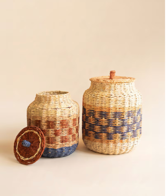 Two woven basket storage jars with lids on a warm beige background. One jar is shown with its lid leaning beside it, revealing a blue and brown patterned bottom.