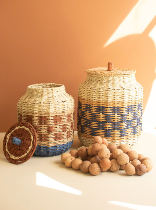 Two woven natural-fiber storage baskets with lids are displayed on a tabletop against a warm terracotta background, with a pile of round wooden beads in front. One basket is shown open with its lid leaning beside it.