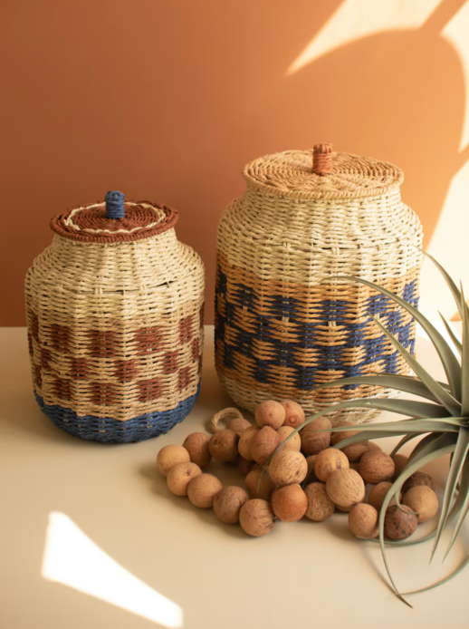 Two woven storage baskets with lids in natural straw tones, one with brown and blue geometric patterns and one with a blue and tan pattern, displayed on a tabletop with cork balls and a plant in front of a warm terracotta background.