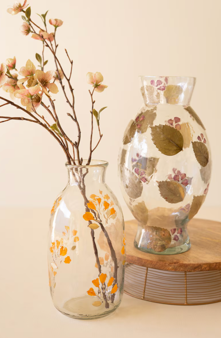 Two clear glass vases decorated with dried botanical elements, one holding delicate pink blossoms and the other standing on a wooden riser. The vases feature painted yellow-orange flowers and green leaves against a warm cream background.