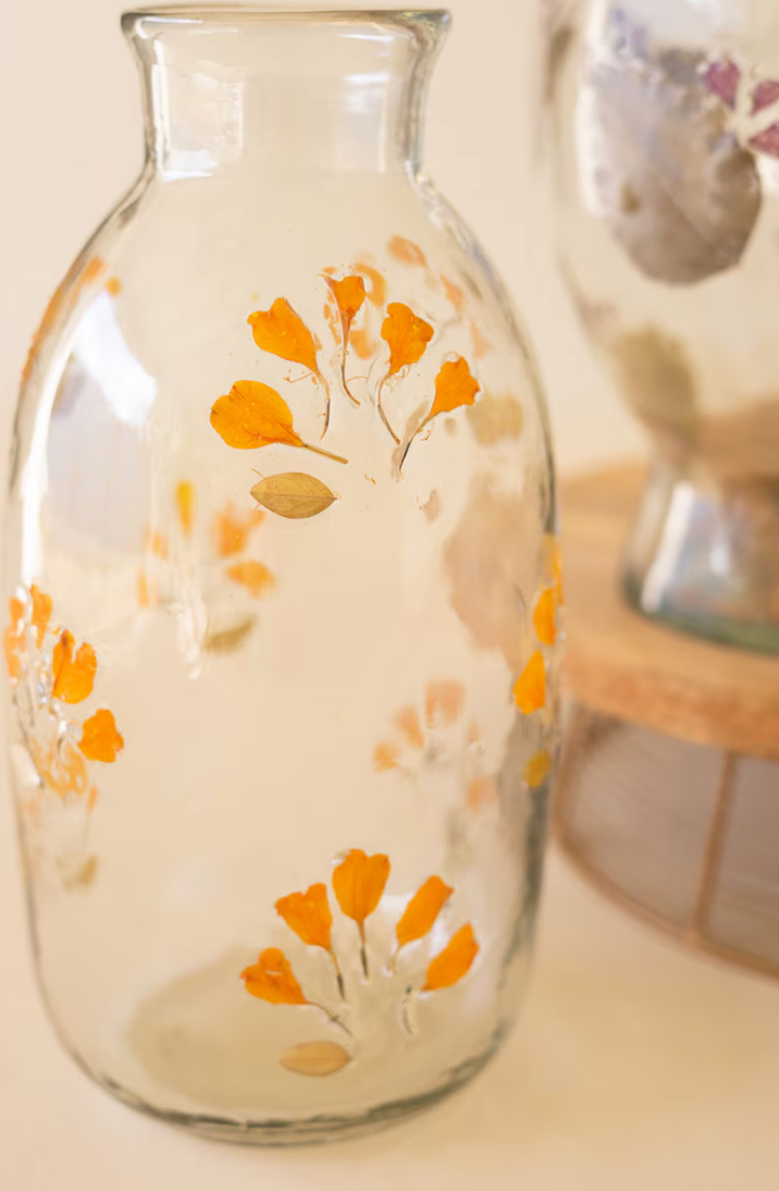 A clear glass bottle with orange and beige botanical leaf patterns painted on the front, shown against a warm cream background with a softly blurred second bottle in the background.