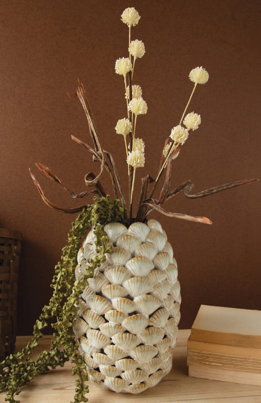 A decorative vase made from stacked seashells in an off-white color, filled with dried branches, cream globe flowers, and trailing green foliage, set on a wooden surface against a brown background.