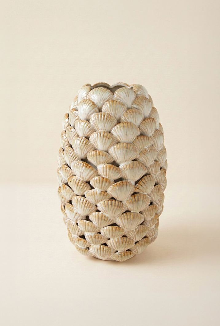 A sculptural vase shaped like a pinecone, featuring a textured surface made of overlapping scallop shell motifs in warm beige and cream tones with subtle brown highlights, set against a solid light beige background.