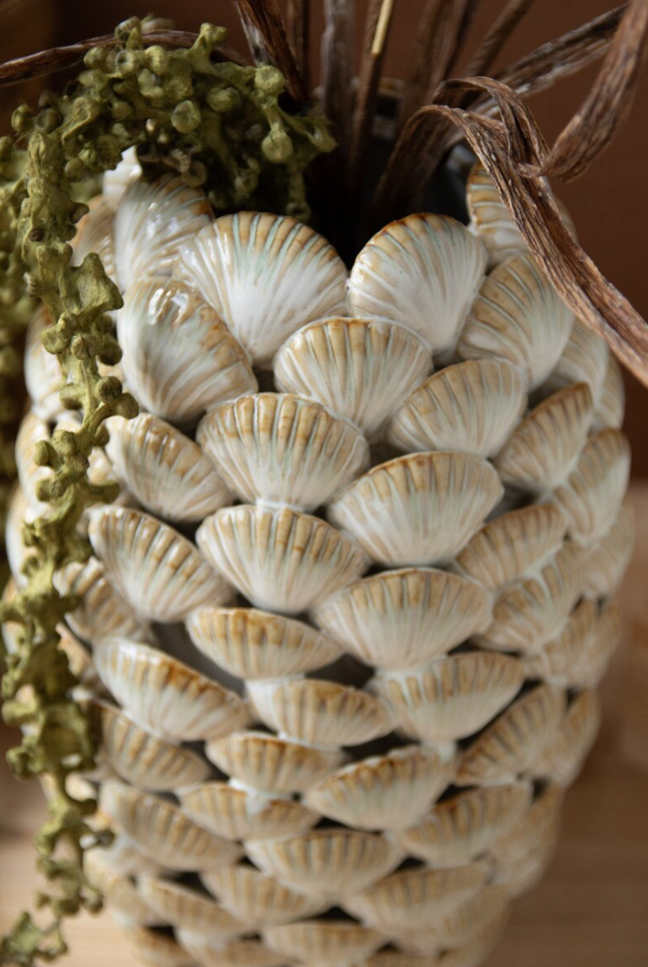A decorative pinecone-shaped vase or container covered in overlapping white and beige seashell patterns, with dried brown stems and greenery emerging from the top, shown in a warm indoor setting.