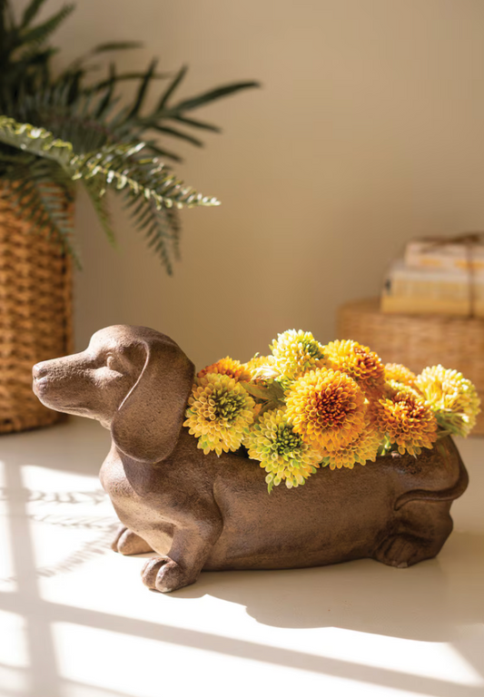 A ceramic dachshund-shaped planter filled with yellow and orange flowers, placed on a tabletop in a warm, sunlit room with a plant and woven basket in the background.