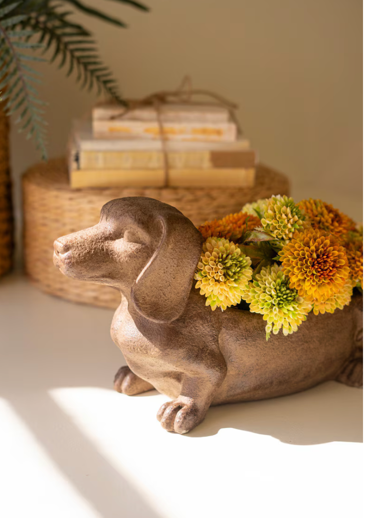A ceramic dachshund-shaped planter in a warm taupe-brown glaze, filled with yellow and orange flowers, set on a white surface with a woven basket and books in the background.