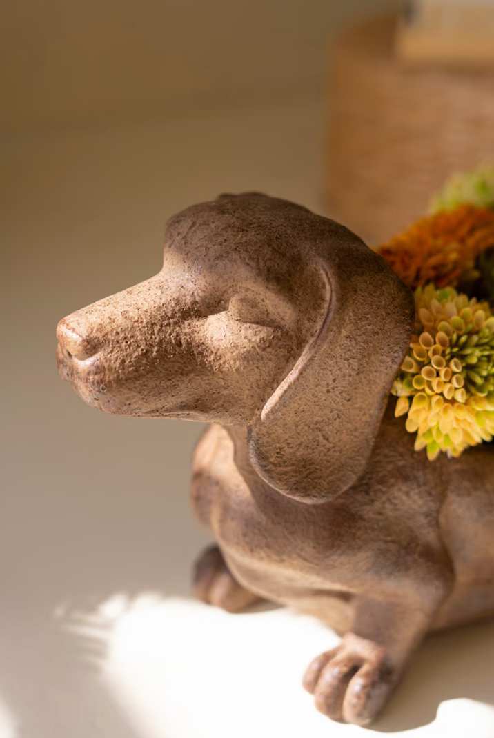 A brown ceramic dachshund figurine with its head turned to the side, decorated with dried orange and yellow flowers on its back, set against a warm beige background.