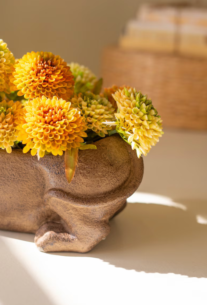 Yellow and orange chrysanthemum flowers arranged in a textured brown ceramic rabbit-shaped planter, set on a warm beige surface with soft sunlight and shadows.
