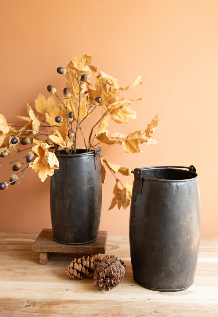 Two rustic black metal canisters, one holding dried yellow oak leaves and berries on a small wooden riser and one empty, styled on a light wood surface against a warm peach background.