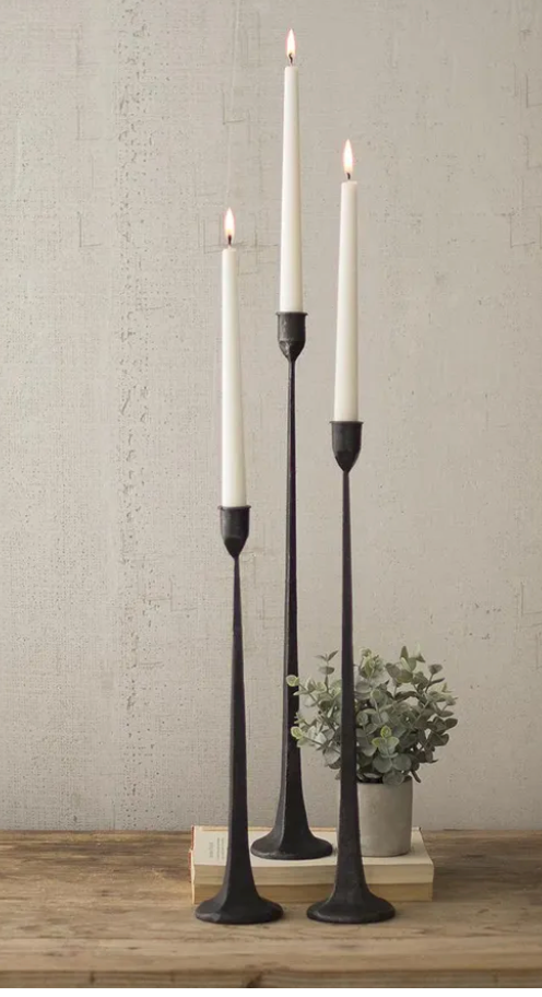 set of 3 thin tall black metal candle holders with white taper candles on each one and the background is white with a wood table. 
