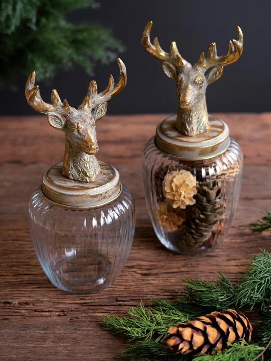 Two clear ribbed glass jars with lids topped by decorative bronze deer heads, displayed on a rustic wooden surface with pine branches and a pinecone.