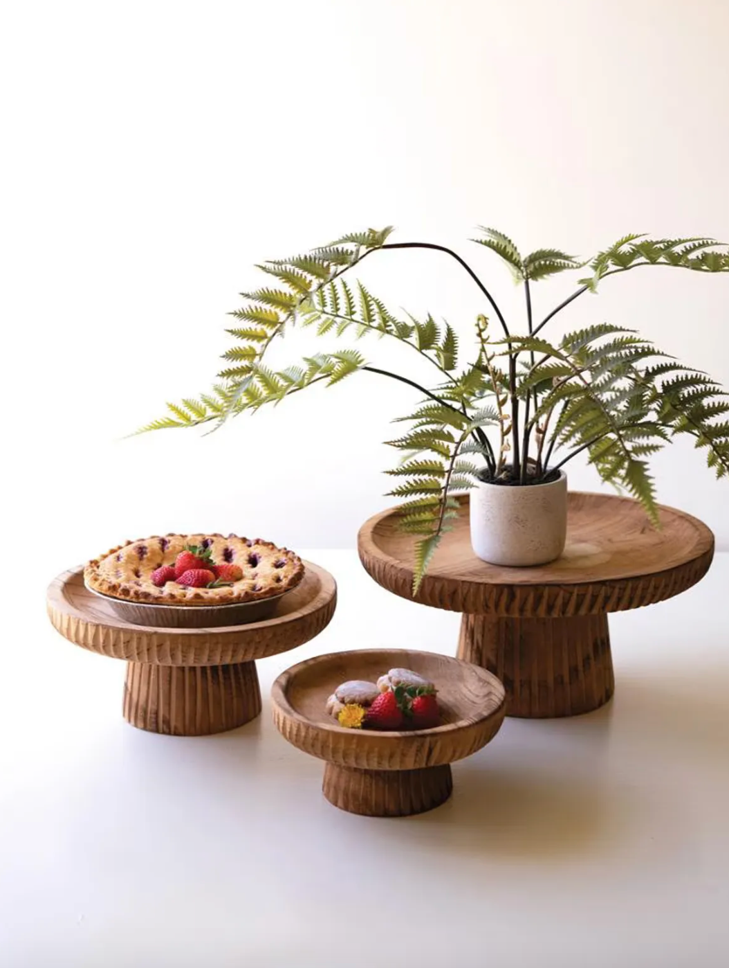Set of three round wooden pedestal stands in graduated sizes, with textured carved edges, styled with a fern, pie, and desserts.