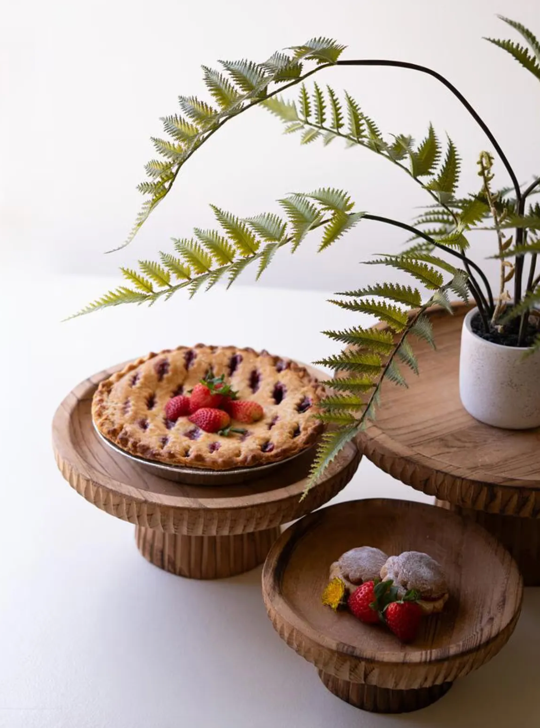 Three wooden display stands holding a berry pie, small desserts with strawberries, and a potted fern.