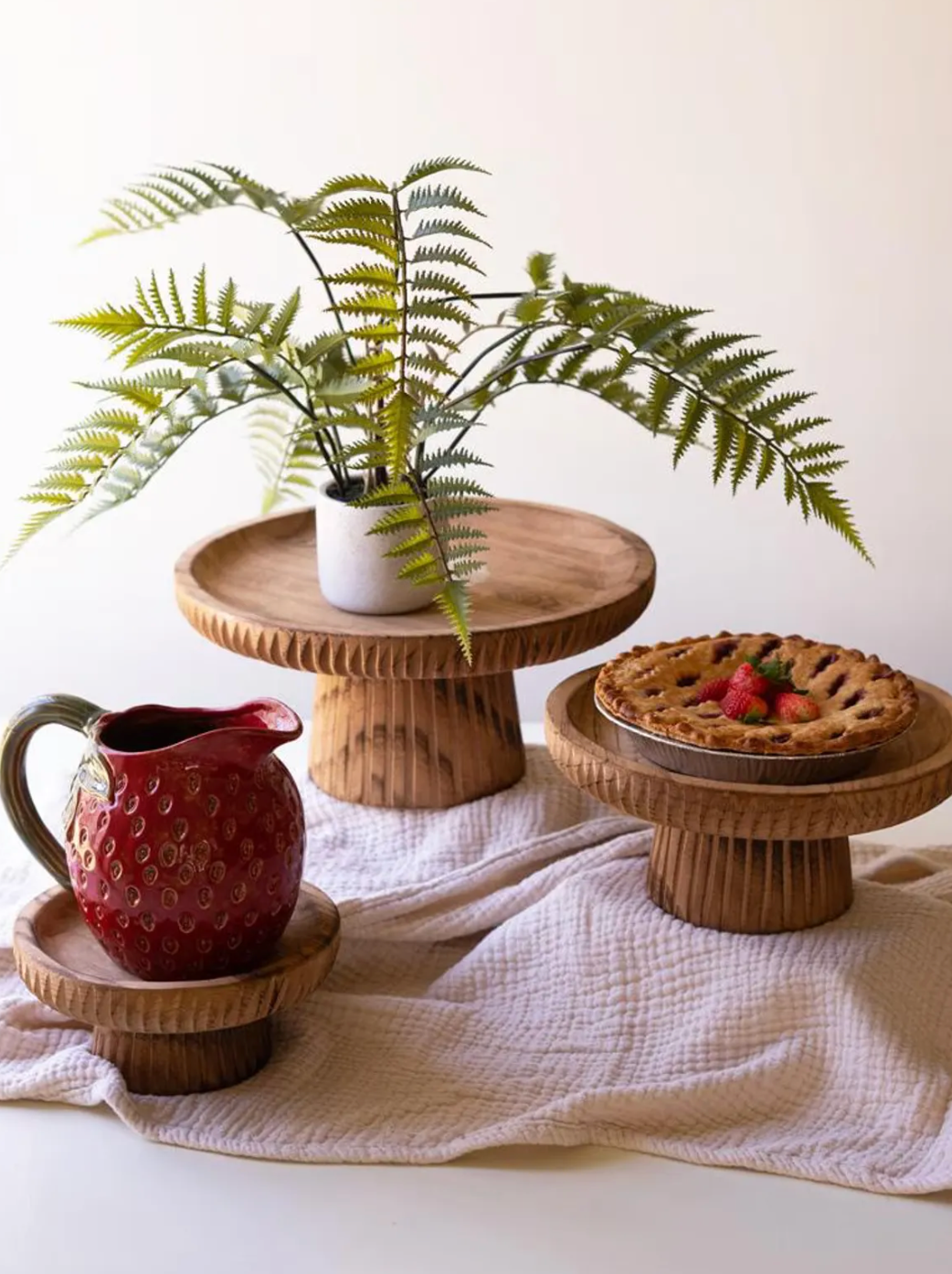 Three carved wooden pedestal stands styled with a potted fern, berry pie, and red strawberry-shaped pitcher on cream fabric.
