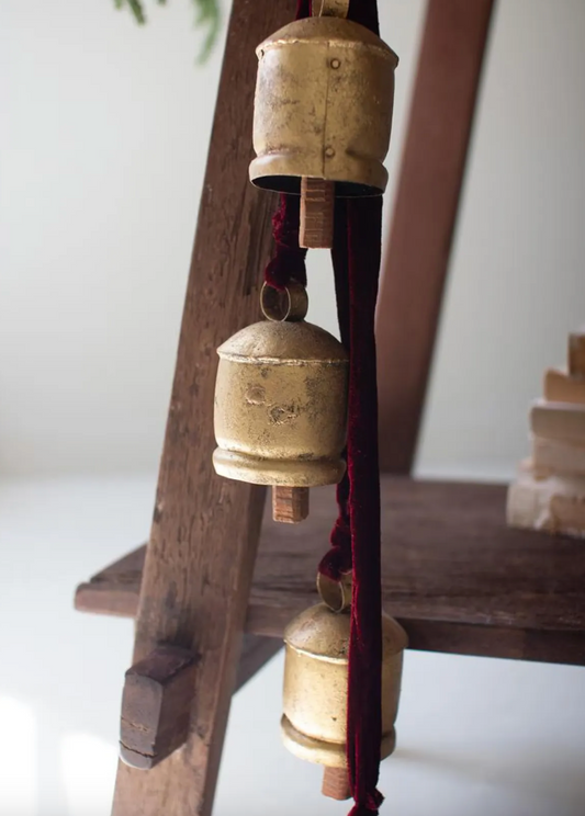 Close-up of distressed gold-tone bells hanging from maroon velvet ribbon beside a rustic wooden ladder.