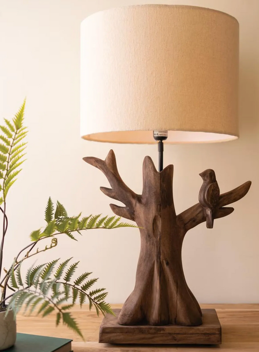 Mango wood tree and bird table lamp with a cream fabric shade, styled on a wooden table beside fern fronds.