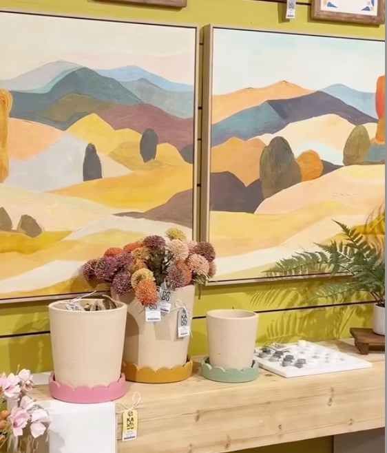 Two framed landscape paintings with plants and a wooden table in the foreground.