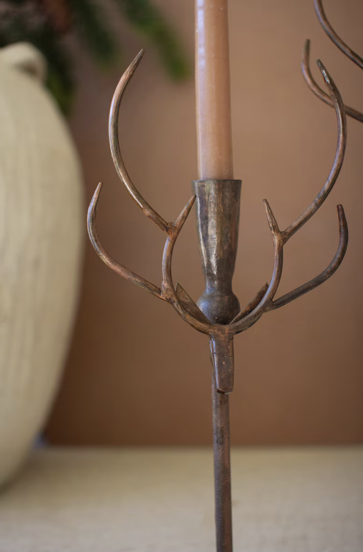 A close-up of a rustic metal deer head candle holder with multiple antler-like branches, holding a beige pillar candle, set against a warm brown background.