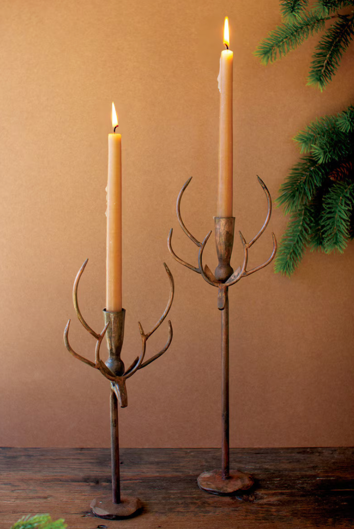 Two rustic metal candlestick holders shaped like antlers, each holding a lit taper candle, set on a wooden surface against a warm brown wall with evergreen branches in the corner.