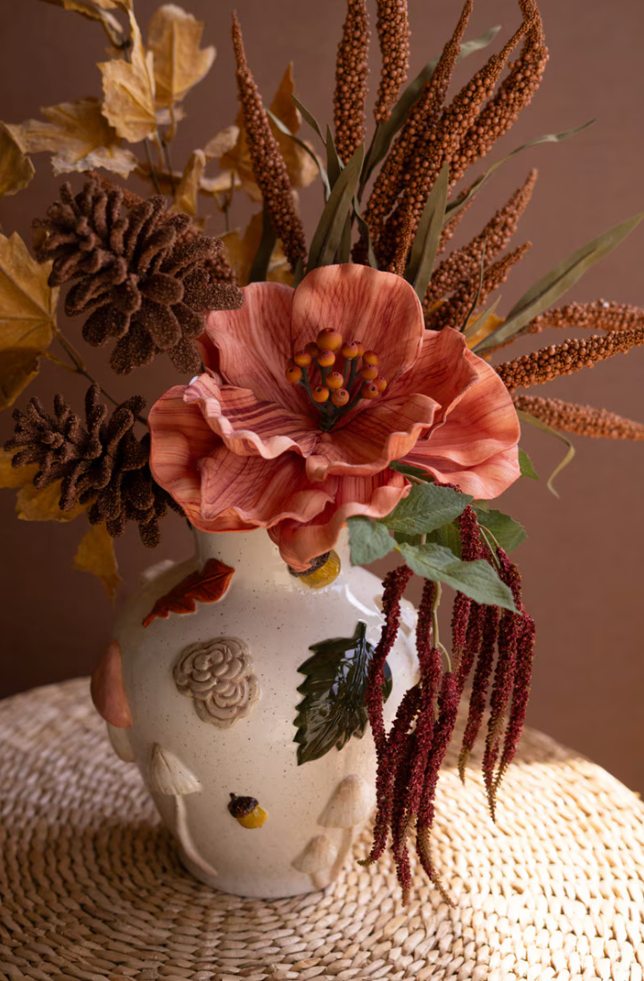 An autumn-themed floral arrangement in a white ceramic vase with hand-painted botanical details, featuring a large peach-pink flower, pinecones, dried brown seed pods, and green leaves, set on a woven placemat against a warm brown background.