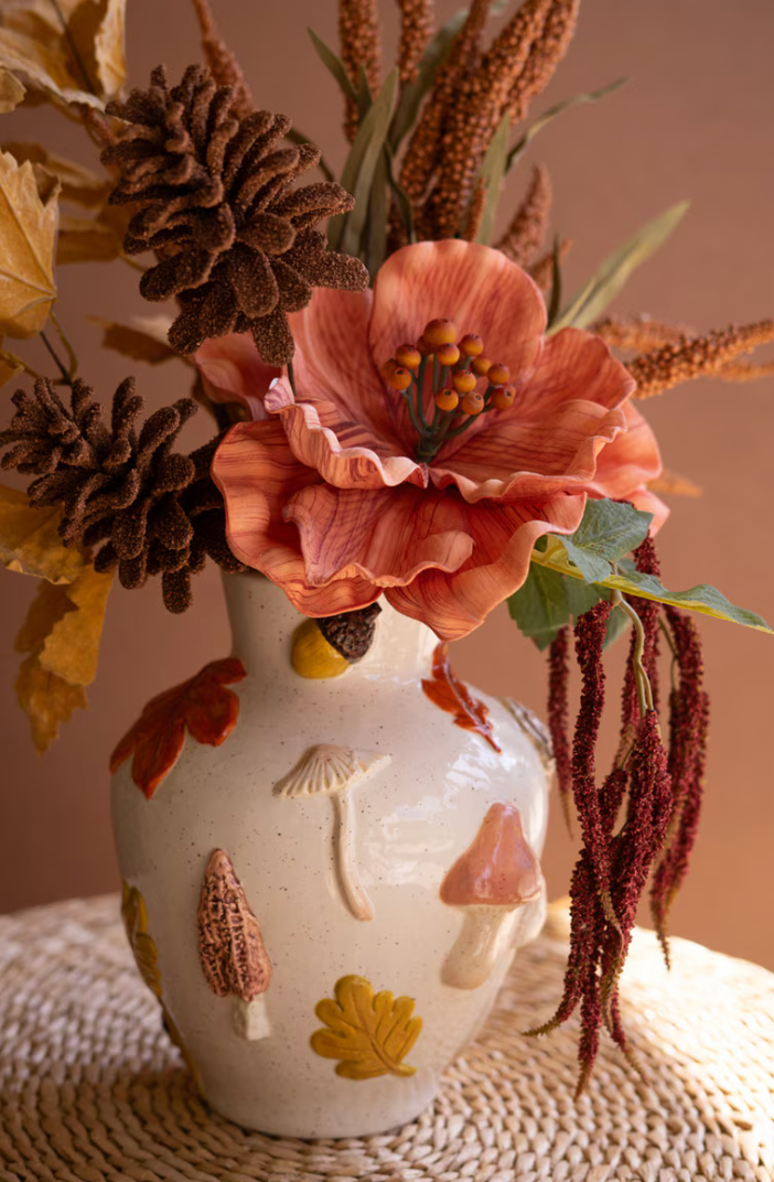 An off-white ceramic vase decorated with raised mushrooms, acorns, and autumn leaves, filled with a dried floral arrangement featuring a large peach lily, pinecones, and warm brown botanicals, set on a woven placemat against a soft beige background.