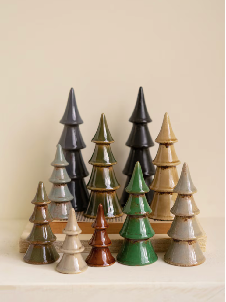 A set of glazed ceramic Christmas tree ornaments in various colors including green, black, brown, beige, and gray, arranged on a wooden stand against a warm beige background.