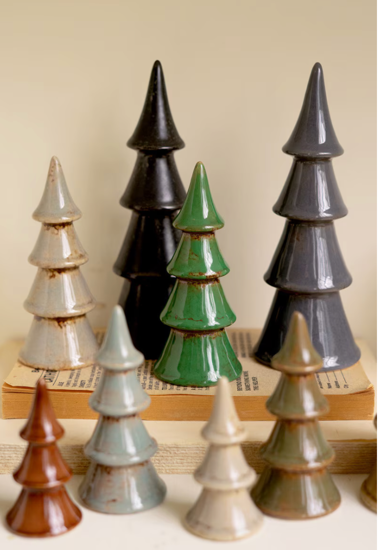 A set of glossy ceramic Christmas tree ornaments in various colors including green, black, gray, brown, beige, and light blue, arranged on top of and in front of vintage books against a warm beige background.