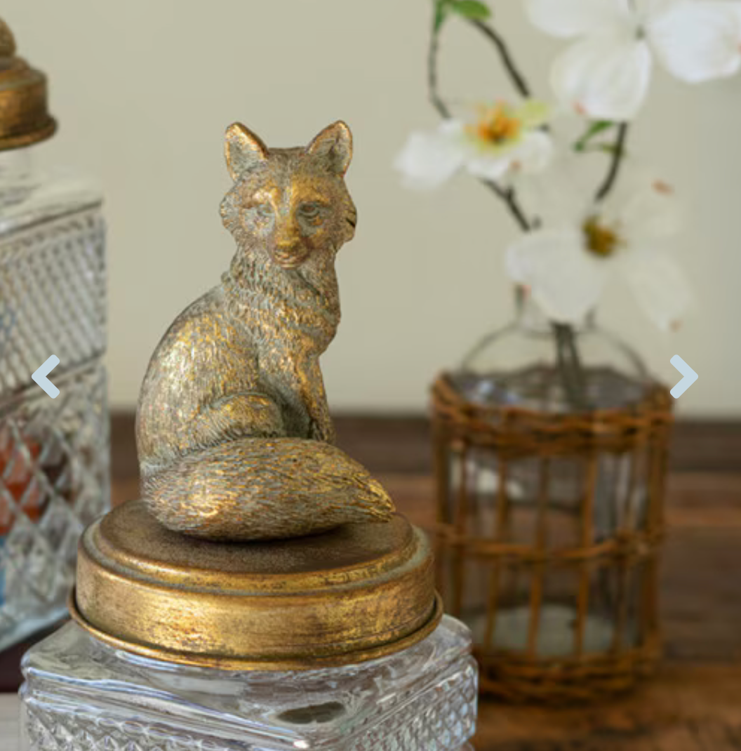 A small metallic fox figurine with a gold and silver textured finish, sitting on a round gold-toned lid atop a clear glass jar, styled on a wooden surface with blurred decorative vases in the background.