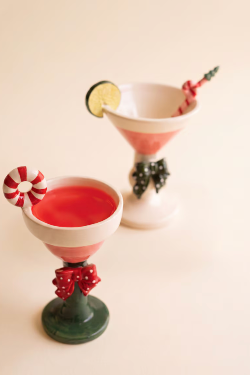 Two festive cocktail-shaped glasses on a warm cream background. The front glass is dark green with a red polka-dot bow and filled with red liquid, garnished with a candy cane.