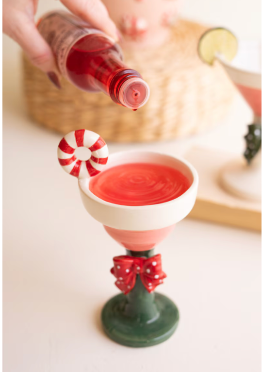 A festive holiday-themed cocktail glass filled with red drink, decorated with a candy cane garnish and a red bow on the stem, while a hand pours a red liquid from a bottle in a softly lit setting.