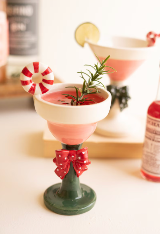 A festive holiday-themed cocktail glass decoration or miniature prop featuring a pink drink, green stem base, red bow, and candy cane garnish, set against a softly blurred background with other drink props.