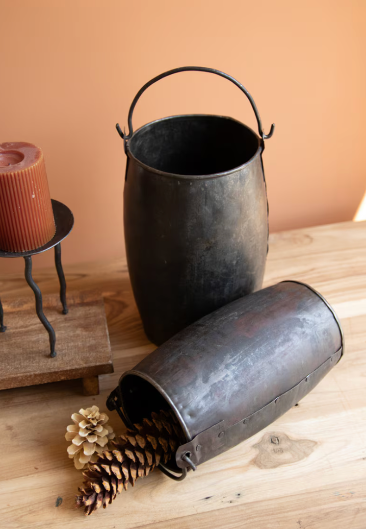 Two rustic dark metal bucket-style containers with handles, one standing upright and one lying on its side, displayed on a wooden surface with pinecones and a candle in the background.