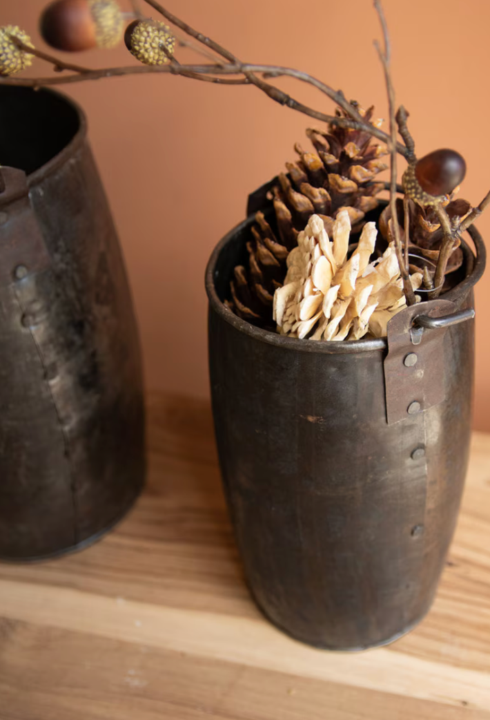 A rustic dark metal bucket-style vase filled with dried pinecones, acorns, and small branches, set on a wooden surface against a warm orange-brown background. Another similar vase is partially visible on the left.