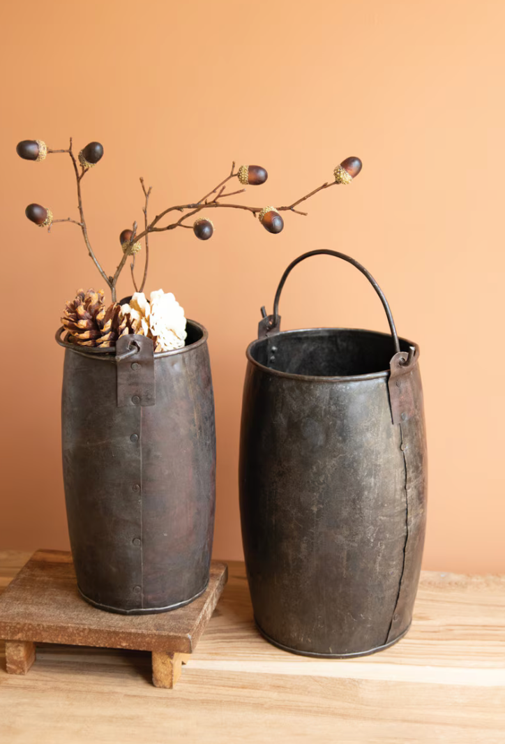 Two rustic dark metal bucket-style vases with handles, one empty and one holding dried branches, pinecones, and a white shell, set on a wooden surface against a warm peach background.