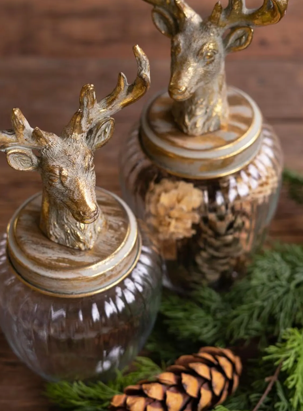 Two decorative glass jars with gold deer head lids, styled with pinecones and evergreen branches on a rustic wooden surface.