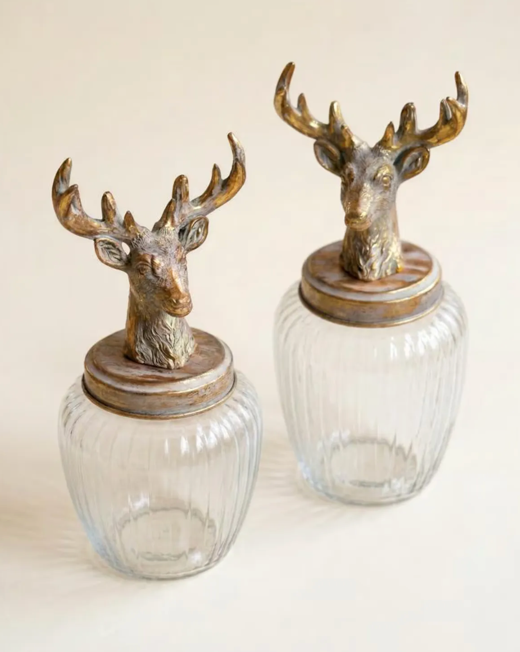 Two ribbed clear glass jars with round lids topped by decorative stag heads in an antique bronze finish, photographed against a plain light beige background.