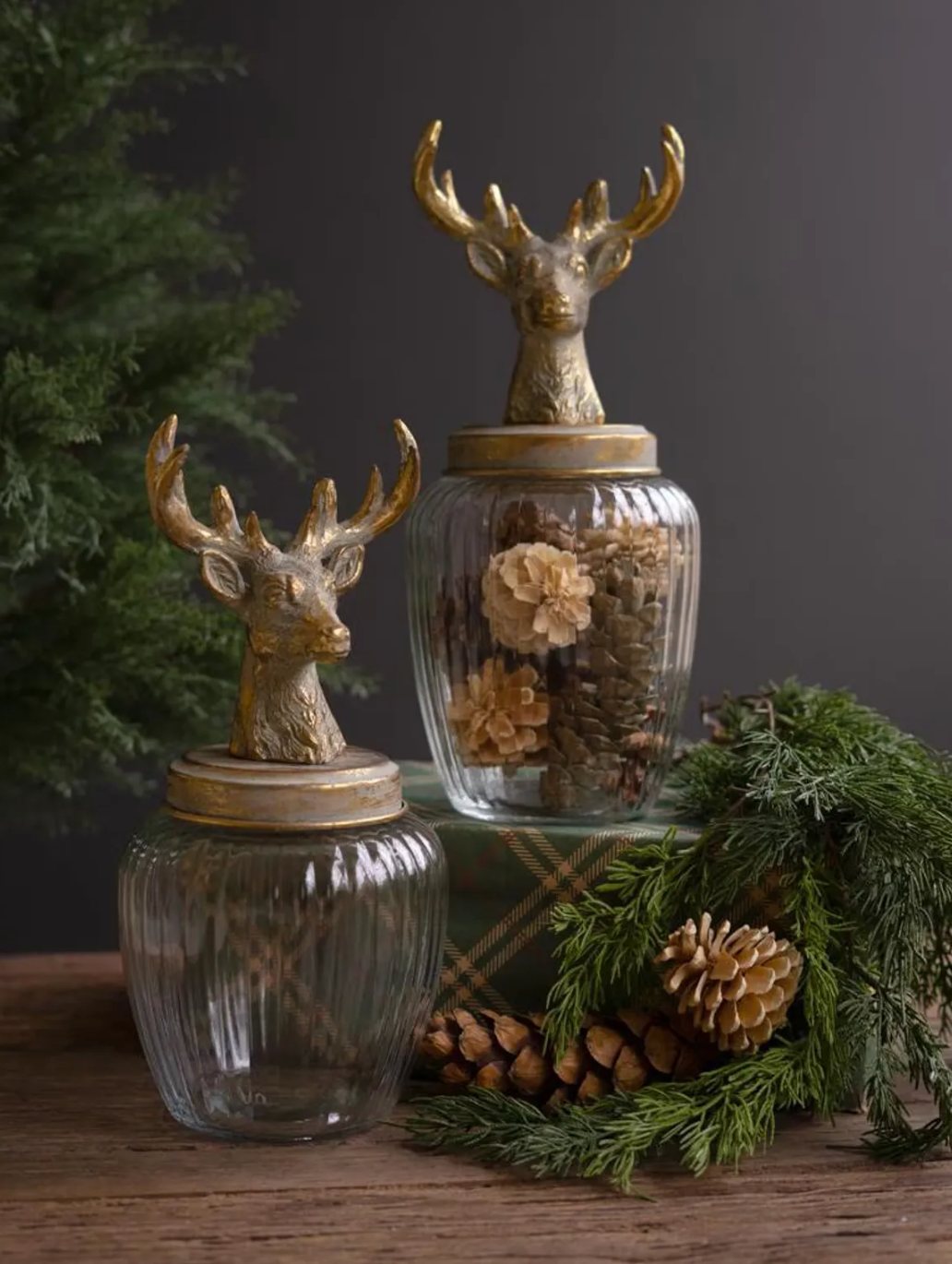 Two decorative glass storage jars with gold deer head lids, styled on a wooden surface with pinecones, evergreen branches, and a plaid gift box against a dark gray background.