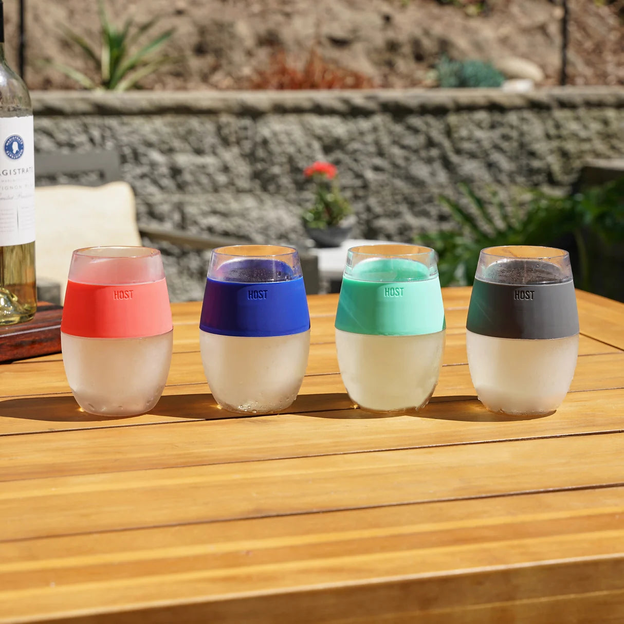 Four colorful stemless wine glasses on a wooden table outdoors.