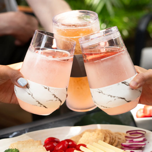 Three clear plastic cups with white marble-patterned silicone sleeves are being clinked together in a toast, filled with pink and orange beverages. Hands are visible holding the cups, and a plate of food is blurred in the foreground, indicating a lifestyle setting.
