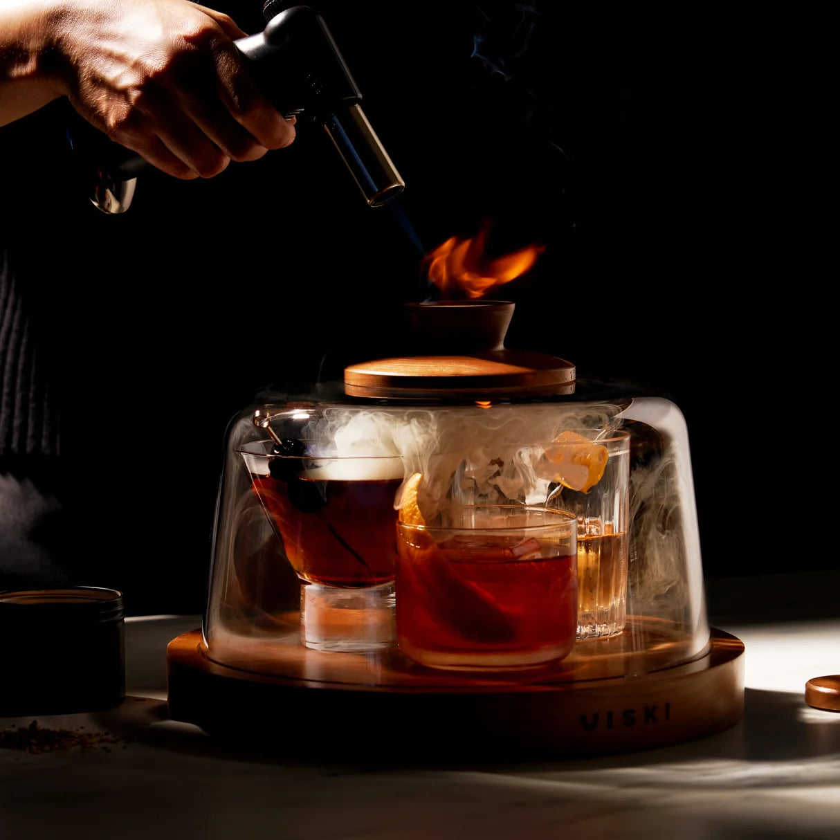 Person using a blowtorch to heat a glass container with a drink inside, on a dark background.