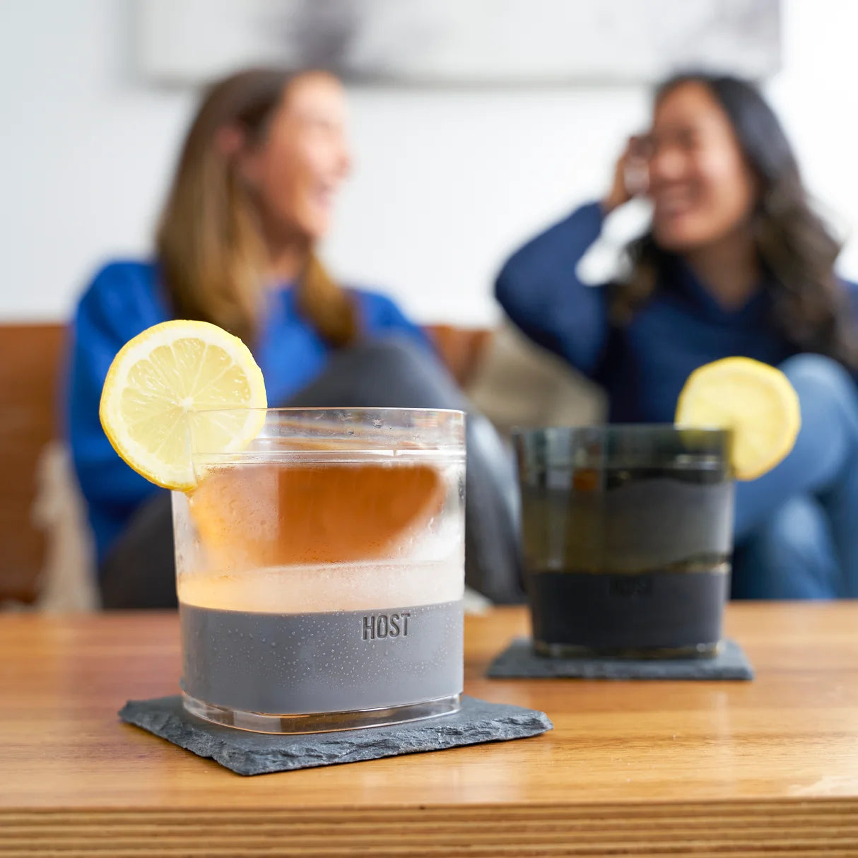 Two glasses with layered drinks and lemon slices on a table, with two people in the background.
