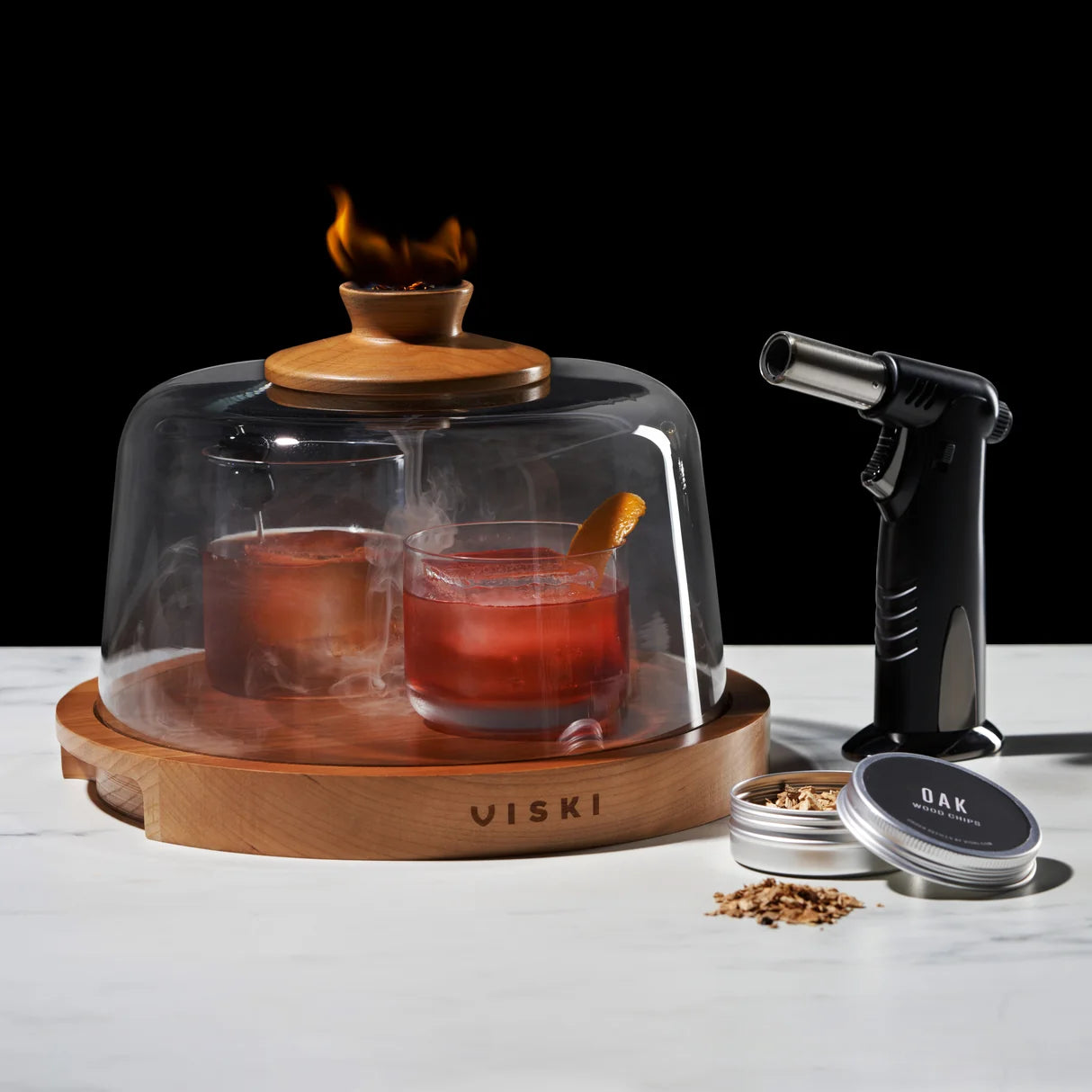 A Viski branded cocktail smoking set on a white marble surface against a black background, featuring a glass dome over two smoking cocktails, a wooden burner torch, and spice containers.