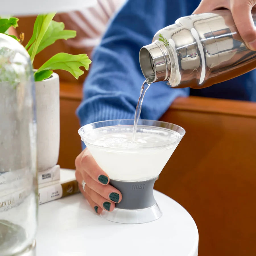 Person pouring a drink into a cocktail glass with a silicone sleeve on a table.